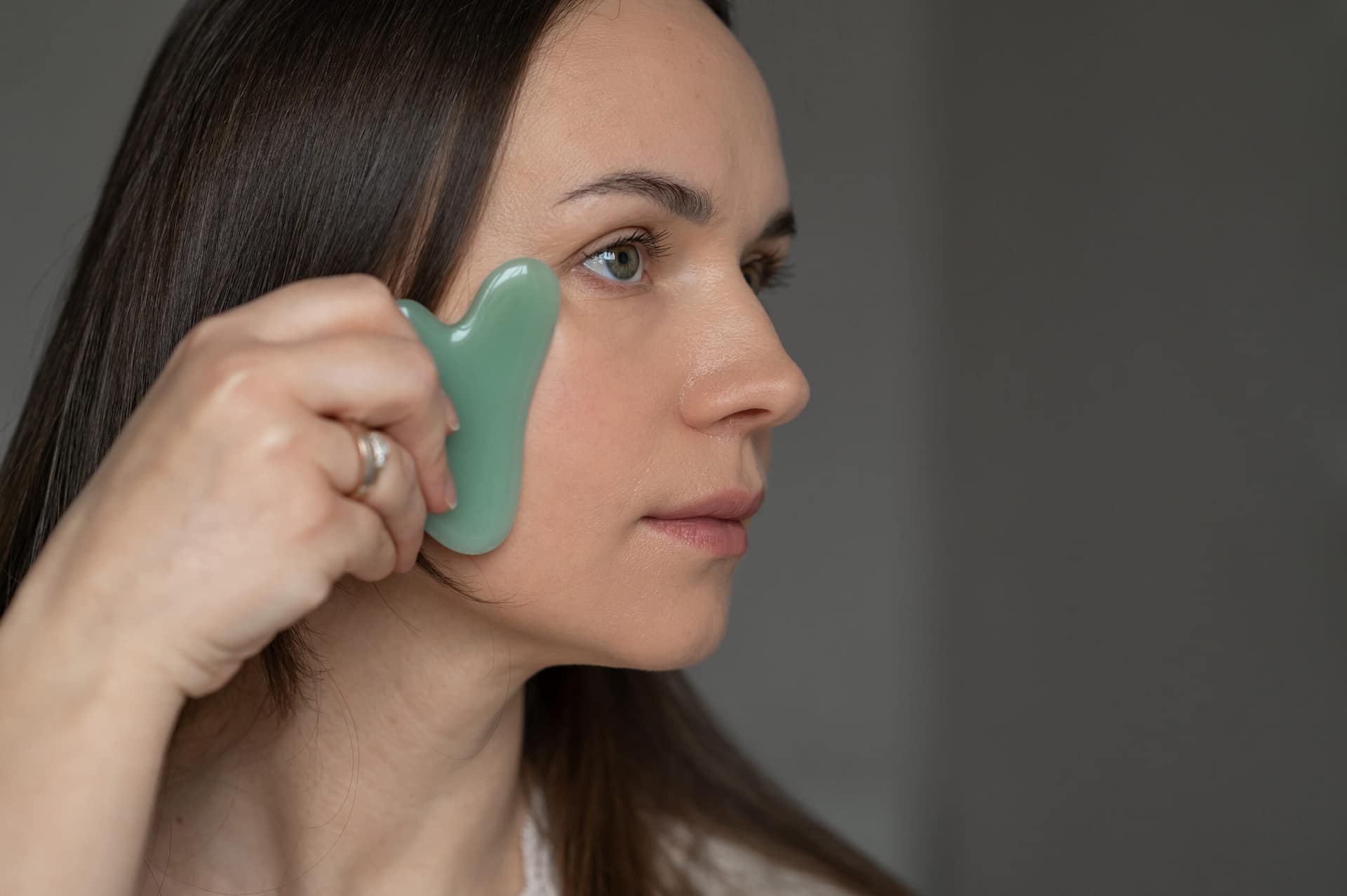 Young woman using a green gua sha scraper for facial massage, improving skin circulation and relaxation. Home skincare and self-care routine for beauty and wellness.